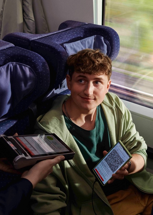 Ein junger Mann in einem Zug zeigt einer Zugbegleiterin einen QR-Code auf seinem Handy. Der Zugbegleiter scannt den QR-Code.